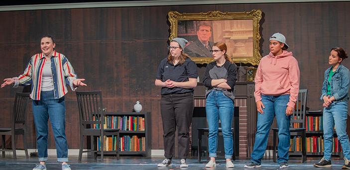 5名演员站在舞台上，背景是壁炉和书柜. 一个演员与其他演员分开，说话和做手势，而其他人在旁边看着.